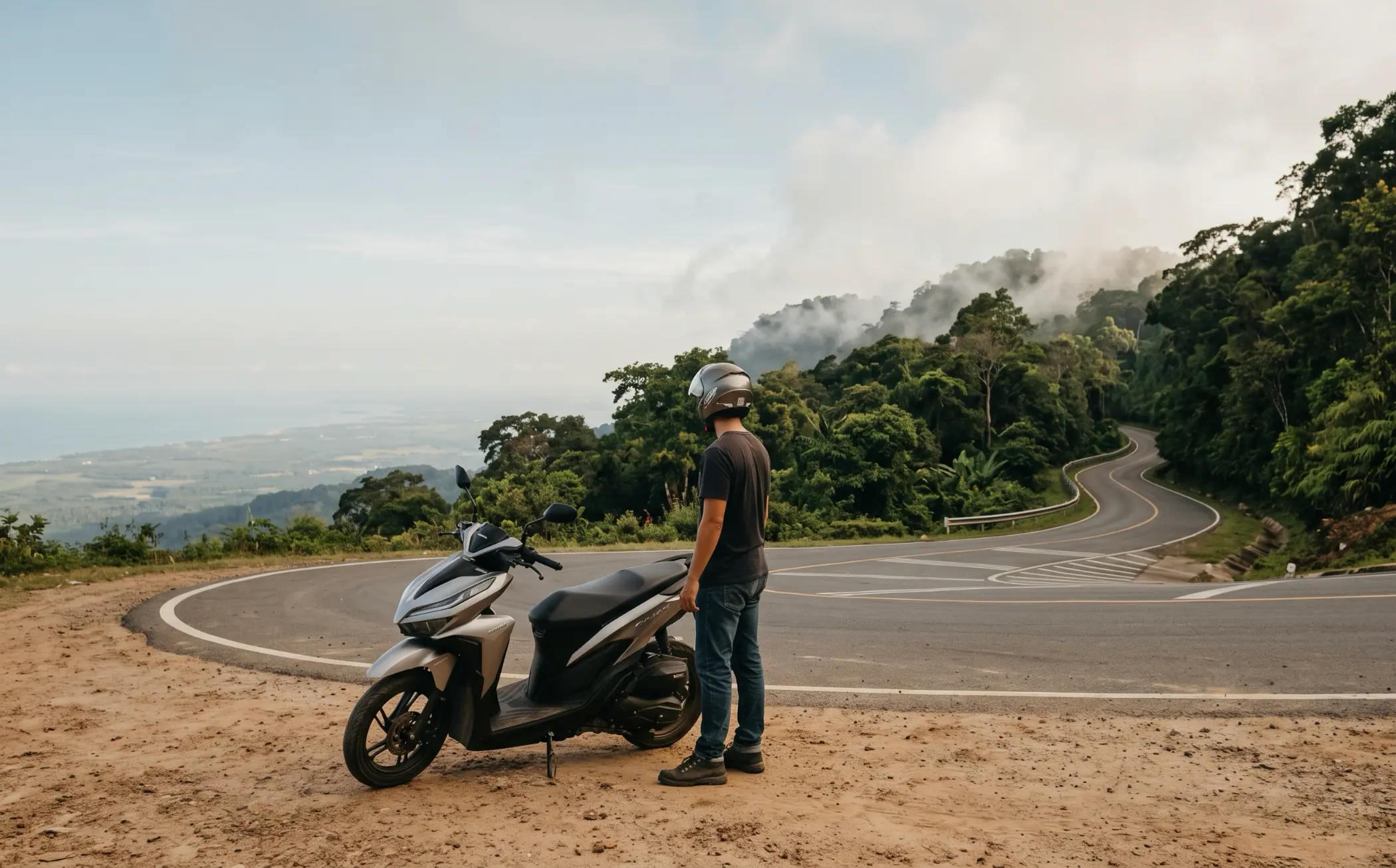 A rider standing beside a parked Honda Click at a hairpin on the Bokor Mountain road, looking out over the coastal plain and the sea far below, cloud lifting off the forested ridge