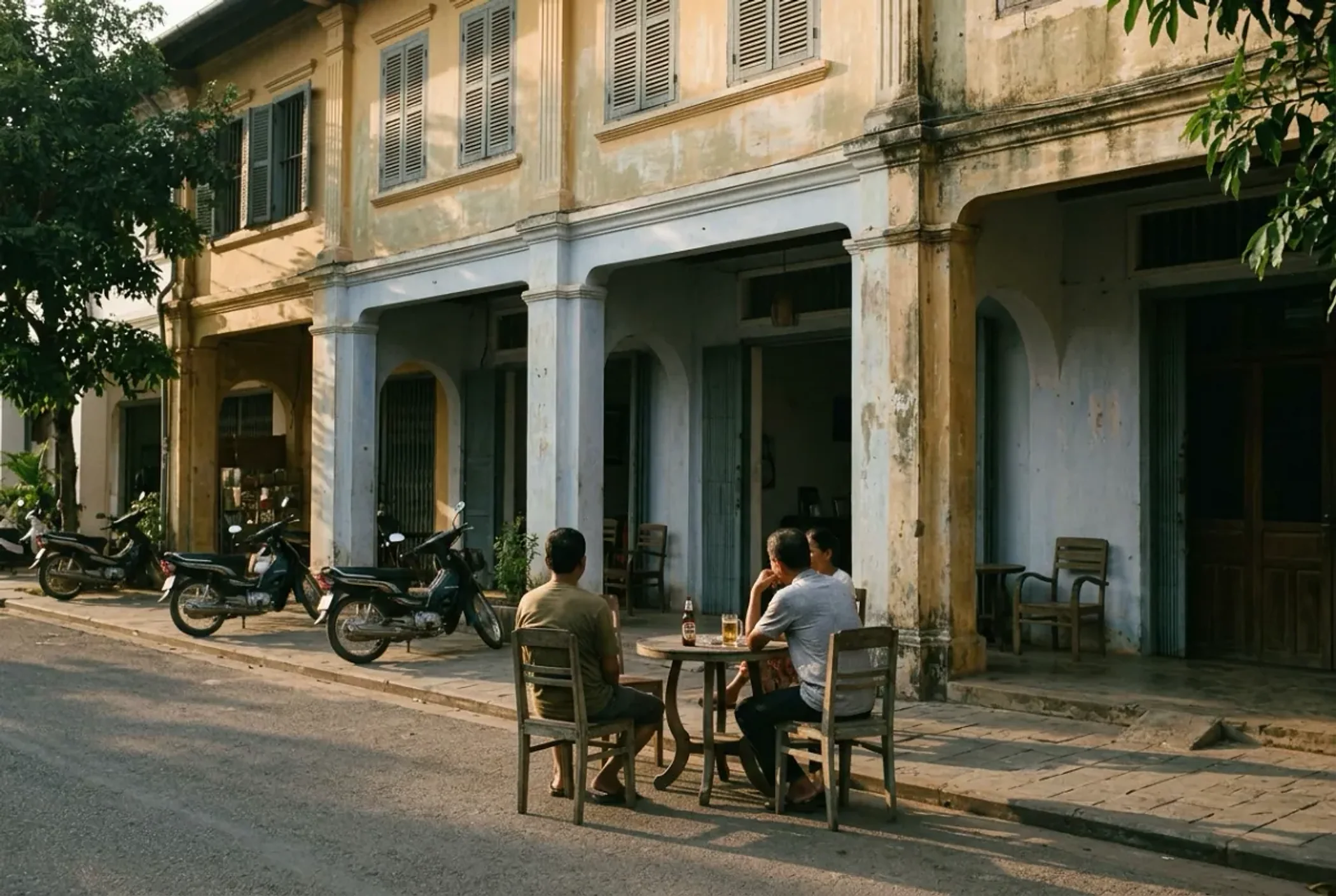 Three people at a wooden table outside a weathered French colonial shopfront in Kampot, late afternoon, motorbikes parked along the kerb