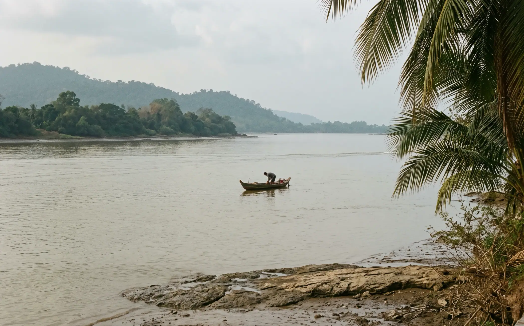 A small wooden fishing boat on the wide Praek Tuek Chhu at Kampot, one figure bent over the nets, wooded hills hazy on the far bank and tide-exposed mud and rock in the foreground