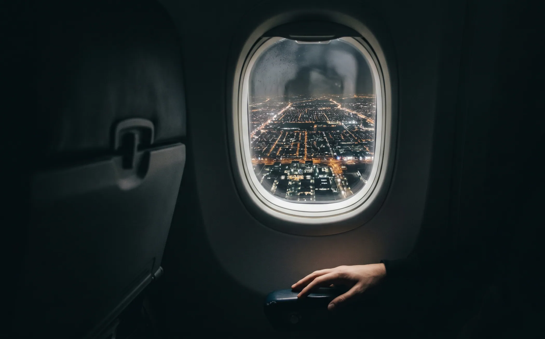 An aircraft window seat at night, a city of lights below, a hand on the armrest - someone in transit between lives