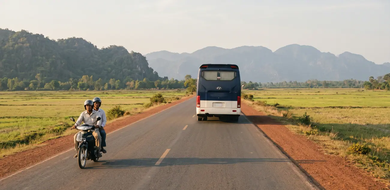 National Road 3 near Kampot in late afternoon: a coach seen from behind on a straight two-lane road through rice paddies, a scooter with two riders alongside, limestone hills on the horizon