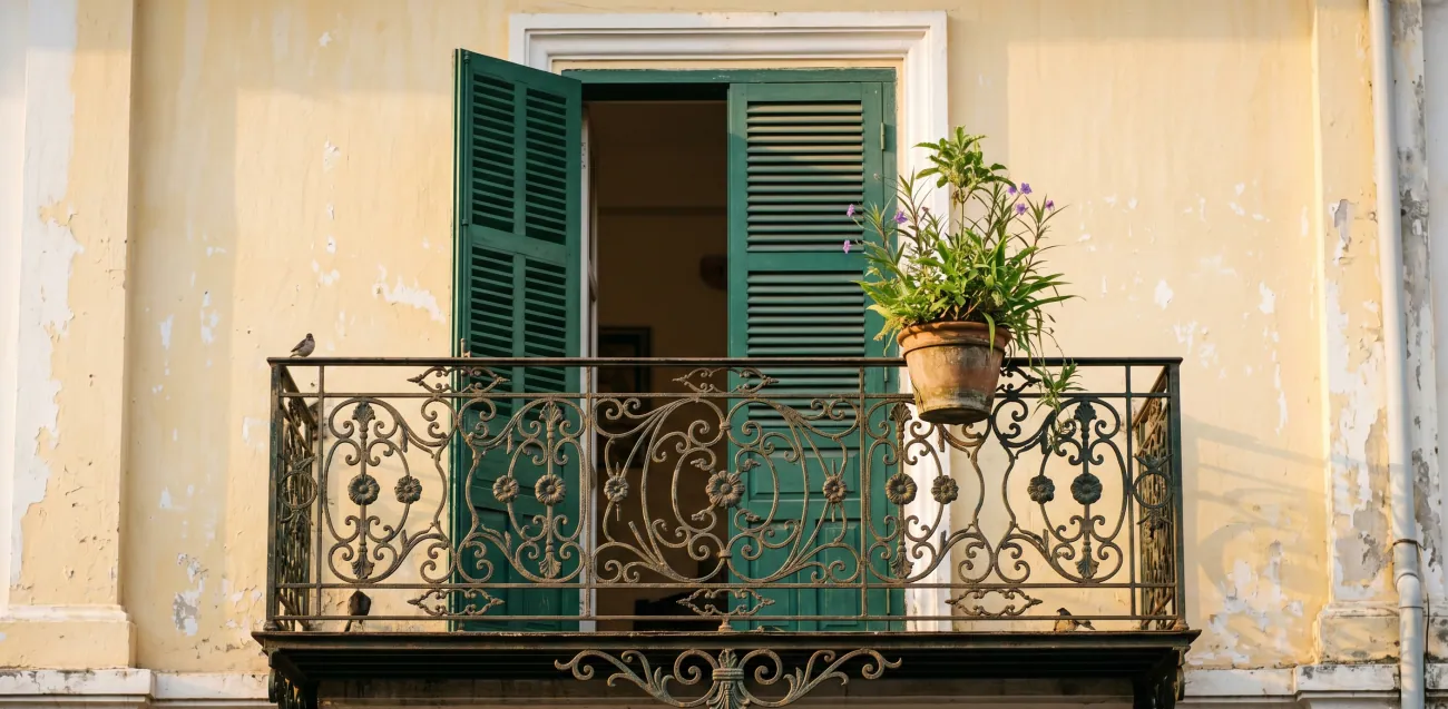 A shophouse facade detail in Kampot's old quarter - a first-floor balcony with iron railings, faded pale yellow wall, a louvred shutter half-open, a potted plant on the railing