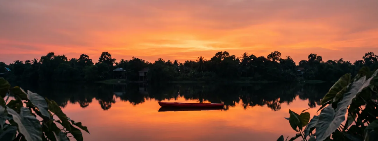 The Praek Tuek Chhu at 5:30pm, sky turning gold, water mirror-flat, a single red kayak in the mid-distance