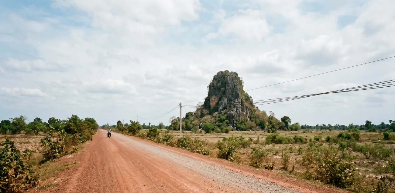 A rural road in flat southern Cambodia leading toward a dramatic limestone karst outcrop in the distance, a single motorbike on the road